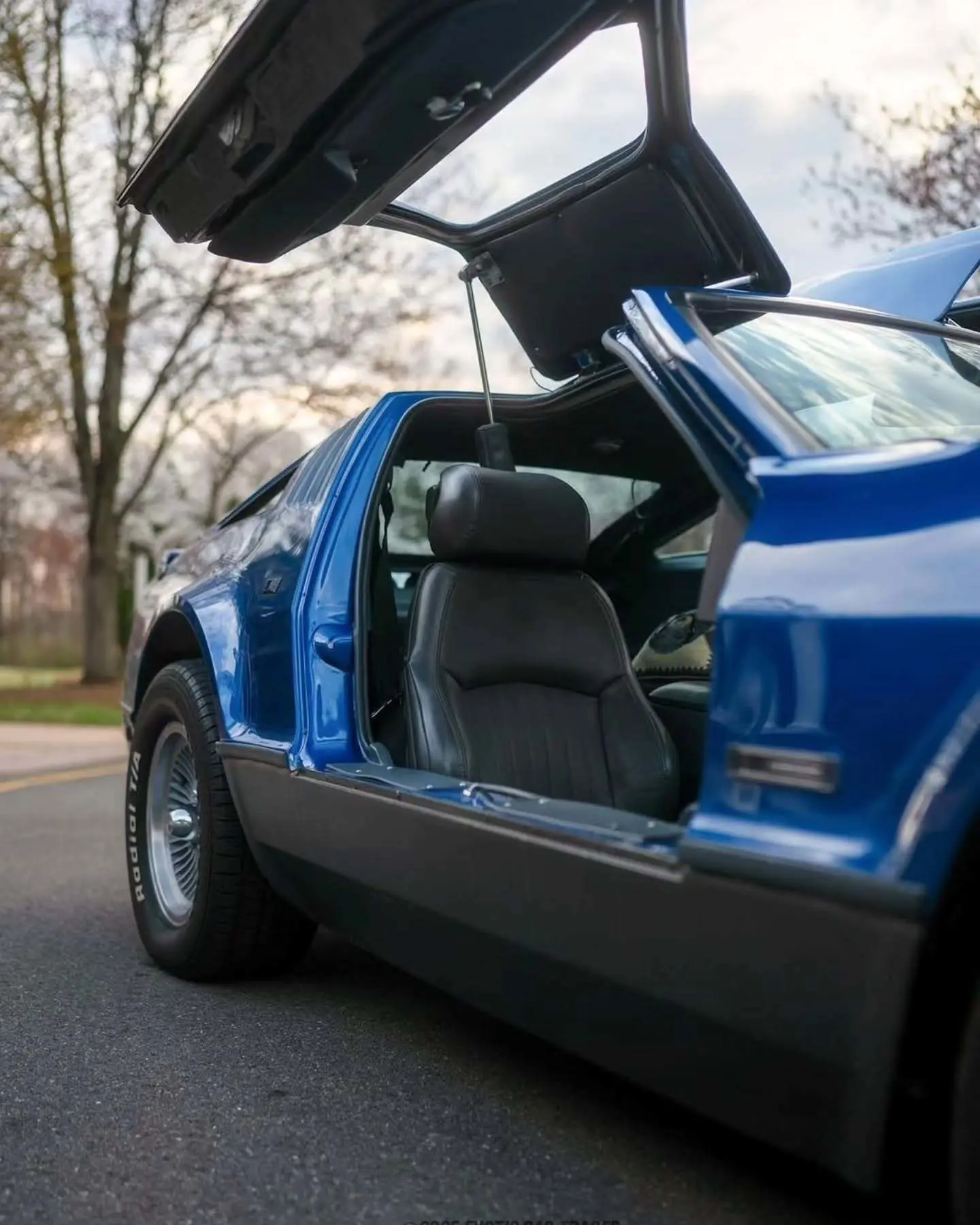 نمای جانبی خودروی کلاسیک بریکلین SV-1 با درهای باز شده رو به بالا و بدنه آکریلیک نارنجی رنگ که طراحی ایمن و آیرودینامیک آن را نشان می‌دهد.